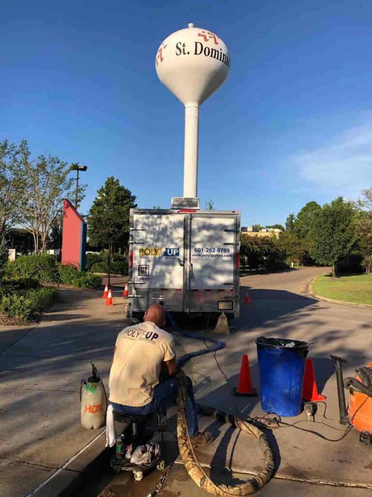 A worker operates equipment near a Poly Up truck, with hoses and cones set up on a street